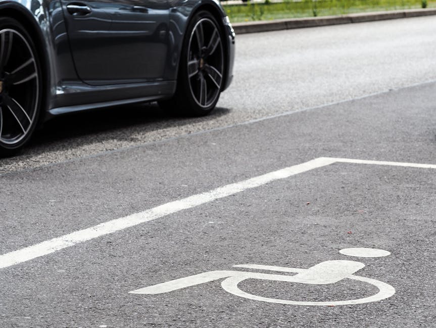 A close-up view of a black modern car parked in a designated parking space on an asphalt surface, with visible black alloy wheels and part of the vehicle's side profile. The parking space features white painted lines and a white wheelchair accessible symbol on the ground, indicating it is a disabled parking bay. The asphalt appears clean and well-maintained, with a textured surface. The setting suggests a residential or commercial parking area, possibly near a property requiring house removal or relocation services. The image is well-lit, capturing the essential details of the vehicle and parking markings, which are relevant for visual context in a house removals service webpage from Man with Van Elmers End, focusing on parking solutions and logistics for furniture transport and home relocation.