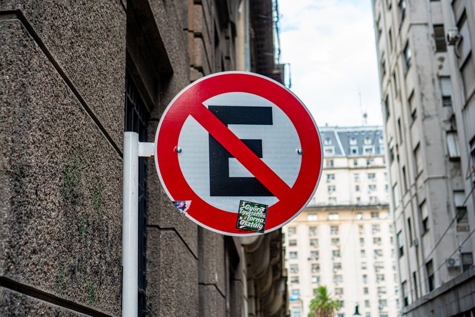 A round traffic sign mounted on a metal pole attached to a textured stone wall, featuring a black letter 'E' with a red diagonal slash across it, indicating no parking. The sign is situated outdoors in an urban environment with tall, multi-storey buildings in the background, some with decorative architectural details and windows. The sky is partly cloudy, and a small green sticker with text is also affixed to the lower part of the sign. This signage is relevant for home relocation and furniture transport services by Man with Van Elmers End, as it highlights parking restrictions that may impact moving or moving-related logistics in Elmers End.