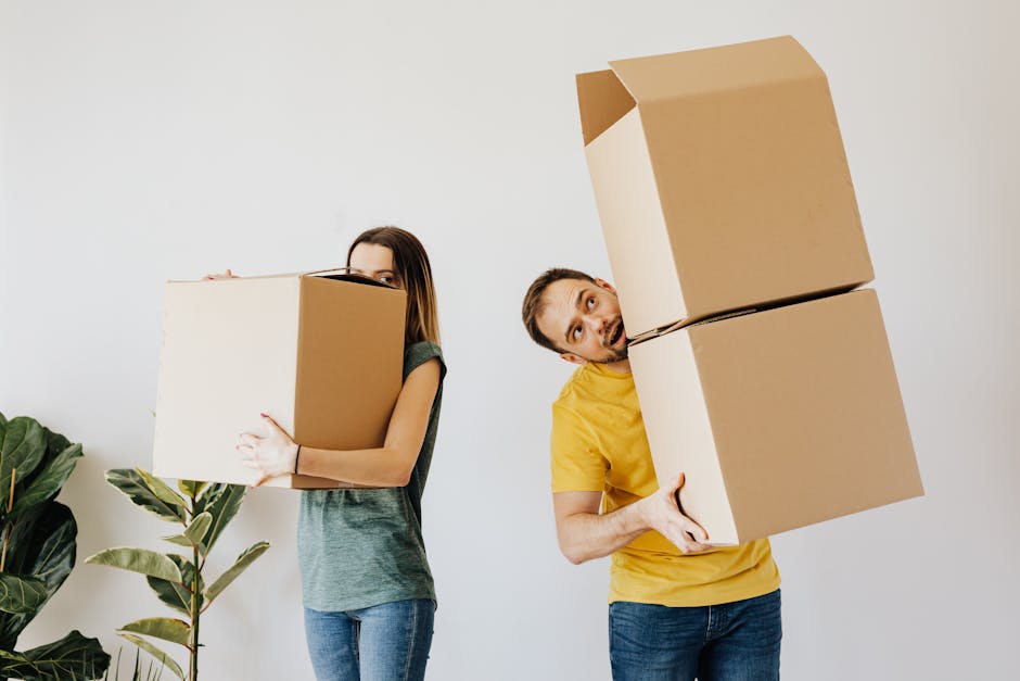 A man and a woman are inside a house, engaged in a furniture transport process as part of a home relocation. The woman, standing on the left, is holding a medium-sized cardboard box close to her chest, wearing a short-sleeved grey top and blue jeans. The man, positioned on the right, is balancing two stacked cardboard boxes, one on top of the other, and is wearing a yellow t-shirt and dark jeans. Both individuals are handling the boxes carefully, indicating a careful packing and moving operation. They are situated inside a bright, well-lit room with plain white walls and a light floor. In the background, part of a green plant with broad leaves is visible, adding a touch of natural décor to the environment. Nearby, a set of gloves and packing materials such as plastic wrap or packing blankets might be present, typical for furniture transport and packing during a house move. The scene captures the logistical aspect of residential removals carried out by [COMPANY_NAME], demonstrating the activity of loading and carrying boxes as part of the moving process.