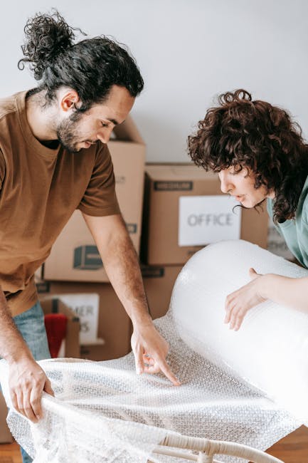 Two individuals, a man and a woman, are engaged in packing or unwrapping a large household item covered by protective bubble wrap, likely in preparation for a house relocation. The man, with long curly hair tied back and a beard, is wearing a brown t-shirt and is pointing at the item while holding the bubble wrap. The woman, with short curly hair, is dressed in a light-colored top and is gently touching the wrapped object, both focused on carefully handling the item. Behind them, there are cardboard boxes labeled for moving, including one marked 'OFFICE,' indicating an organized packing process inside a residential space. They are standing on a wooden floor, with additional packing materials and boxes visible in the background, suggesting a professional move or furniture transport arranged by Man with Van Elmers End. The scene illustrates a careful, systematic approach to packing and loading delicate household goods for a home removal or furniture transport service.