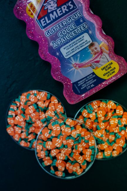 Close-up image showing a pink, glittery container of Elmer's glitter glue with a label featuring a young girl demonstrating its use, placed on a dark surface. Next to the container are three round tin