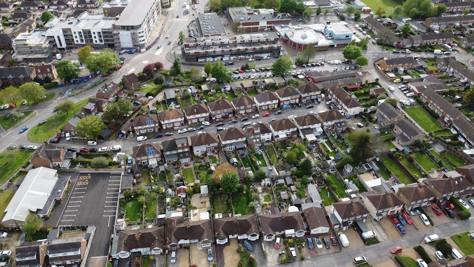 Aerial view of a residential neighbourhood showing multiple terraced houses with small gardens and backyards, some featuring patio furniture, sheds, and decorative plants. Several cars are parked along the street and in driveways, with a few visible on the pavement. A larger commercial or apartment building with a parking lot is positioned at the top of the image. In the foreground, a house is being prepared for relocation, with visible packing materials such as cardboard boxes and plastic wrapping, as well as a moving van from Man with Van Elmers End parked nearby. The scene captures the loading process during a home relocation, with someone seen lifting or carrying boxes towards the vehicle. Green trees and lawns are dispersed throughout the neighbourhood, contributing to the suburban setting, which aligns with house removals and furniture transport services in the context of Bromley Council permits for street-loading at Elmers End.
