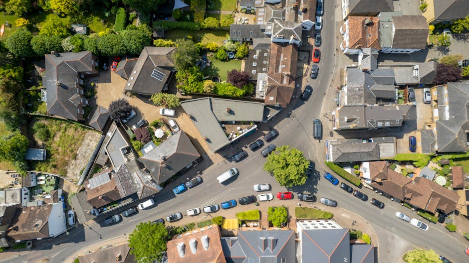 Aerial view of a residential street showing parked cars lining both sides of the road, with a mix of houses and gardens visible. The street is narrow and features a small roundabout with a single tree in the center, around which multiple vehicles are parked or moving. Several houses have visible roofs with solar panels or chimneys, and some gardens contain trees, bushes, and lawn areas. A delivery van is parked near a property, suggesting ongoing home relocation or furniture transport activities. The scene is lit by daylight, with shadows cast by trees and buildings, highlighting the densely built neighborhood typical of house removals and moving services in Elmers End, as managed by Man with Van Elmers End, supporting street-loading permits for household moves.