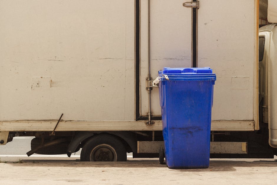 A blue plastic waste bin positioned outside a white delivery van, which is parked on a pavement. The van's side panel is slightly worn with visible dirt and weathering, and the lower section reveals part of the wheel and undercarriage. The waste bin is placed near the van's open rear door, ready for rubbish or bulky waste collection, relevant to house removal or furniture disposal services. Inside, a glimpse of the van’s interior door frame and sliding mechanism is visible, indicating the vehicle is prepared for loading or unloading bulky items, such as furniture or packaging materials. The scene suggests a home relocation process, possibly involving the disposal of unwanted household items like sofas or mattresses, aligning with services provided by Man with Van Elmers End for bulky waste pickups and efficient furniture transport.