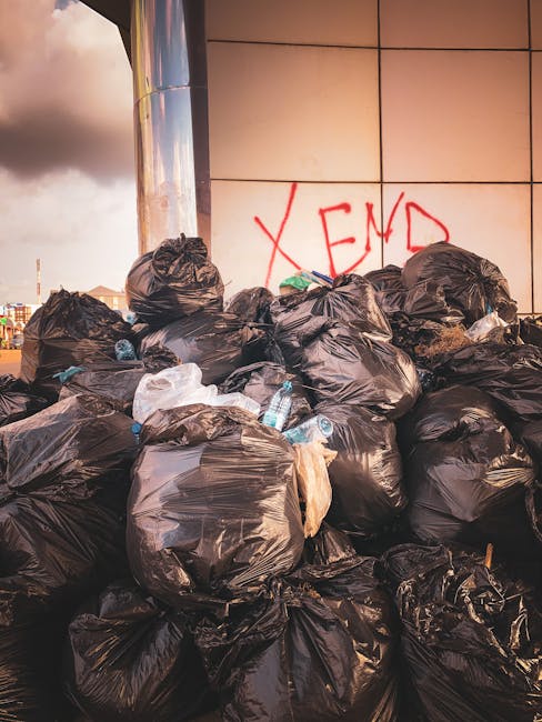A large pile of black rubbish bags filled with household waste, including visible plastic water bottles and packaging, stacked against a tiled wall in an outdoor urban area during daylight. Behind the rubbish bags, there is red graffiti on the wall that reads 'XEND'. The scene appears to be in a public space near a building, with the light of the setting sun casting a warm glow. This image relates to bulky waste disposal and house clearance services, as handled by Man with Van Elmers End, highlighting the process of removing unwanted household items during a home relocation or clearance, with a focus on efficient loading and waste removal.