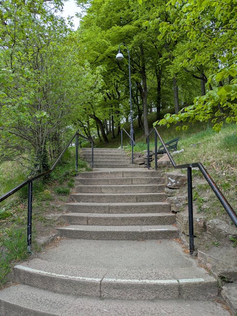 The image depicts a set of outdoor concrete steps leading uphill through a green, leafy environment. The stairs are flanked by metal handrails on both sides and are situated beside a grassy slope with scattered rocks and trees. A lamppost with a white globe light fixture is positioned along the pathway near the top of the stairs, which is surrounded by dense foliage and branches of tall trees, creating a shaded area. At the top of the stairs, there is a small, flat landing that appears to lead into a wooded area. This scene illustrates the typical access route used during house removals or home relocations in residential settings, highlighting the challenges of navigating stairs and access points in the process, as managed by [COMPANY_NAME].