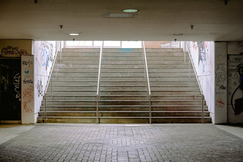 A narrow outdoor staircase composed of concrete steps with visible wear, leading uphill from a cobbled pathway. To the left, a tall brick retaining wall supports the slope, while on the right, a wooden handrail runs alongside a small garden area with bushes and plants. The staircase is shaded by overhanging tree branches and foliage, with dappled sunlight filtering through. In the background, the top of the stairs is partially obscured by trees, suggesting a residential setting. The scene appears to be part of a house move or relocation, and the environment indicates a typical UK suburban area, where [MAN WITH VAN ELMERS END] provides house removals and furniture transport services involving careful handling of packed boxes, furniture, and home appliances to navigate access points such as stairs during a home relocation.