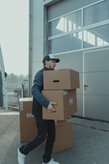 A man wearing a dark grey cap, maroon polo shirt, and red shorts is positioned outdoors on a concrete surface, pushing a black hand trolley loaded with three large cardboard boxes. The boxes are seale