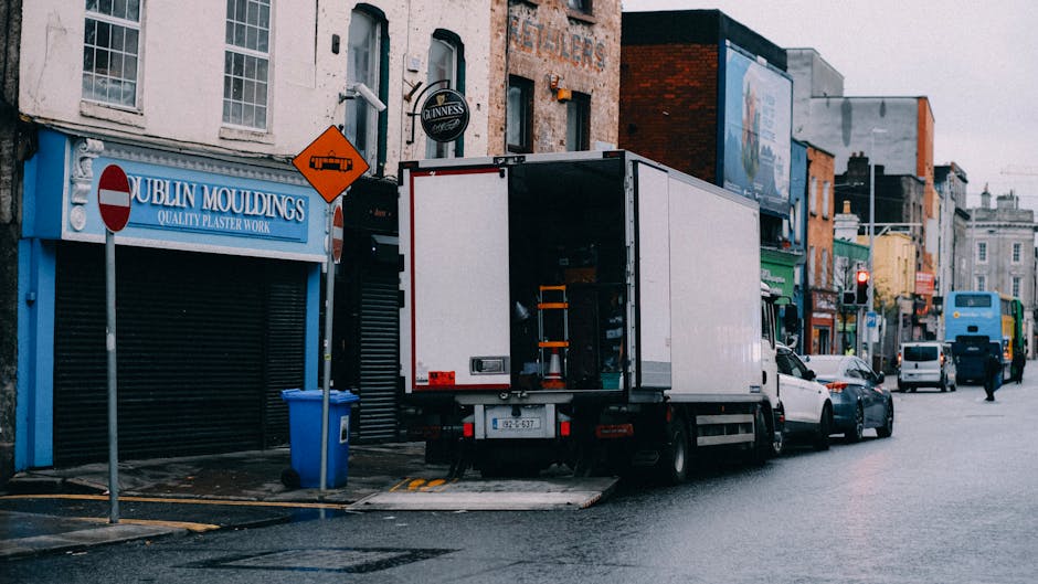 A large green box trailer attached to a commercial vehicle is parked on a city street in front of tall modern office buildings and historic architecture. The trailer is situated near the pavement, wit