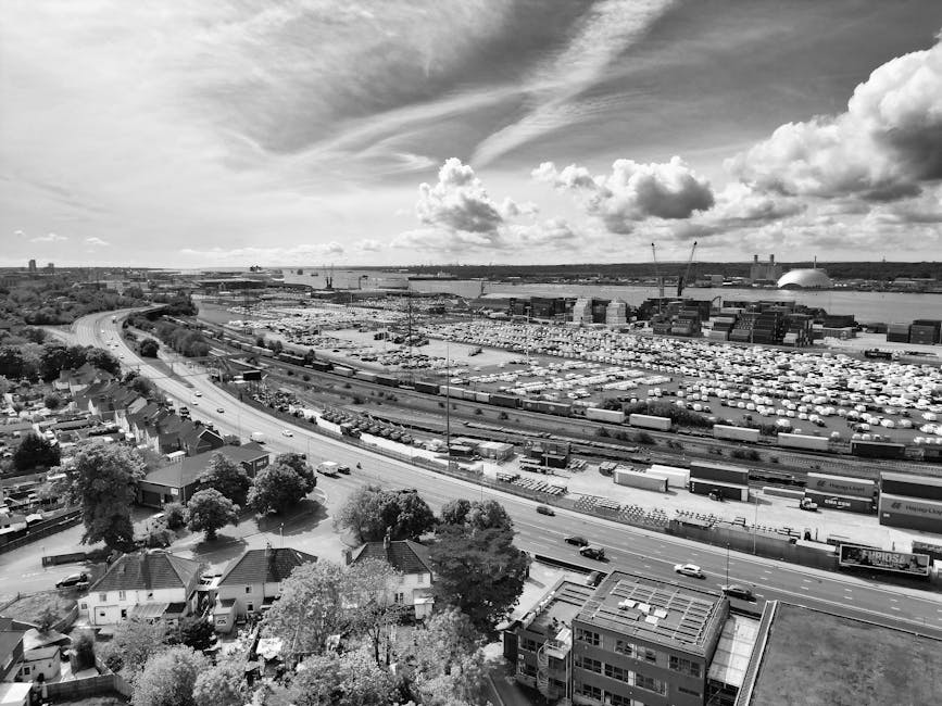 A black and white aerial photograph of a large industrial port area with numerous docked cargo ships and stacked containers, situated along a wide river. In the foreground, there are residential house