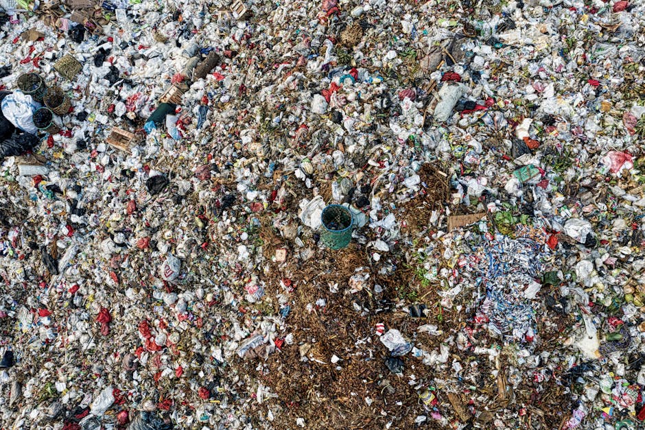 A large area of waste and household rubbish including plastic bottles, cardboard boxes, paper, and discarded packaging materials scattered across the ground outdoors. The waste is a mix of different colours and textures, with some items partially covered by soil and debris. Amidst the rubbish, a small green plastic bucket is visible, along with other scattered objects. The environment appears untidy and unclean, reflecting improper waste disposal. This scene illustrates the importance of proper waste management and waste disposal rules near Elmers End, as part of a home relocation or moving process supported by Man with Van Elmers End, a professional removals service specializing in residential moves and furniture transport.