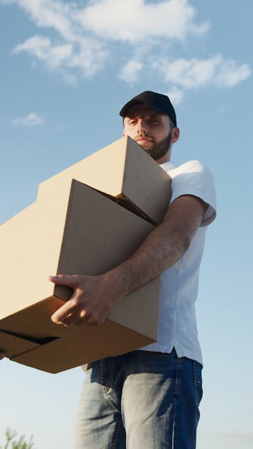 A man wearing a black cap, white t-shirt, and jeans is outdoors under a partly cloudy sky, holding three cardboard moving boxes stacked in his arms. The boxes appear to be of medium size, with some edges slightly worn, and are secured with clear packing tape. The man's expression is neutral as he looks into the distance. In the background, part of a paved driveway or street is visible, suggesting he is involved in house relocation or furniture transport. The scene reflects the process of home moving, with the man ready to load or unload the boxes, supporting professional removals services like those offered by Man with Van Elmers End. The natural lighting and open environment emphasize the practical aspect of packing and moving, integral to reliable and efficient moving logistics.