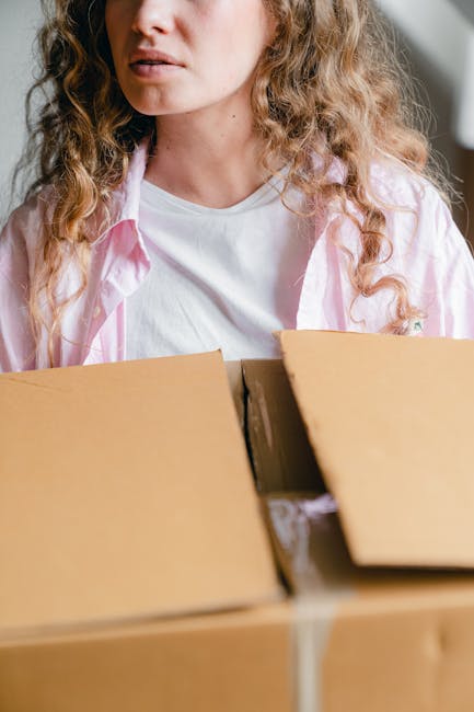Inside a residential room with wooden flooring and large windows with grey curtains, several moving boxes made of brown cardboard are visible, some placed on the windowsill and others on the floor. One box is being carried or positioned by an individual whose white socked feet are partially visible behind the boxes, indicating a careful handling process during packing or unpacking. The boxes vary in size and are sealed or open, with no visible packing materials such as bubble wrap or packing paper. The natural daylight from the windows illuminates the scene, providing a clear view of the moving process. This image, associated with house removals and furniture transport, highlights the packing and organization involved in home relocation, as managed by Man with Van Elmers End, who offers professional removal services in Elmers End.
